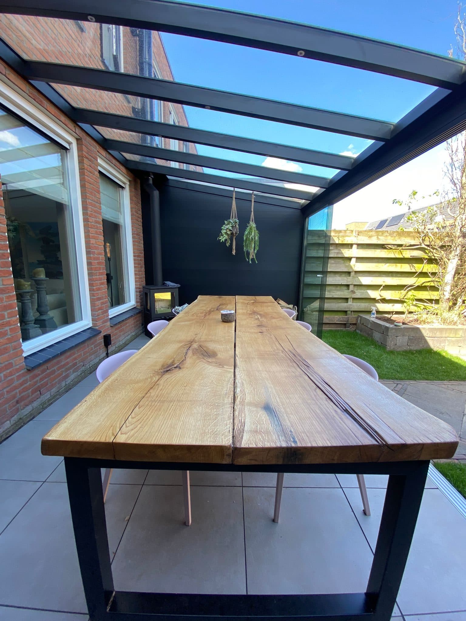 Lange houten tafel onder een zwart glasdak met een dichte zijwand en een houtkachel, van onder het dak gezien