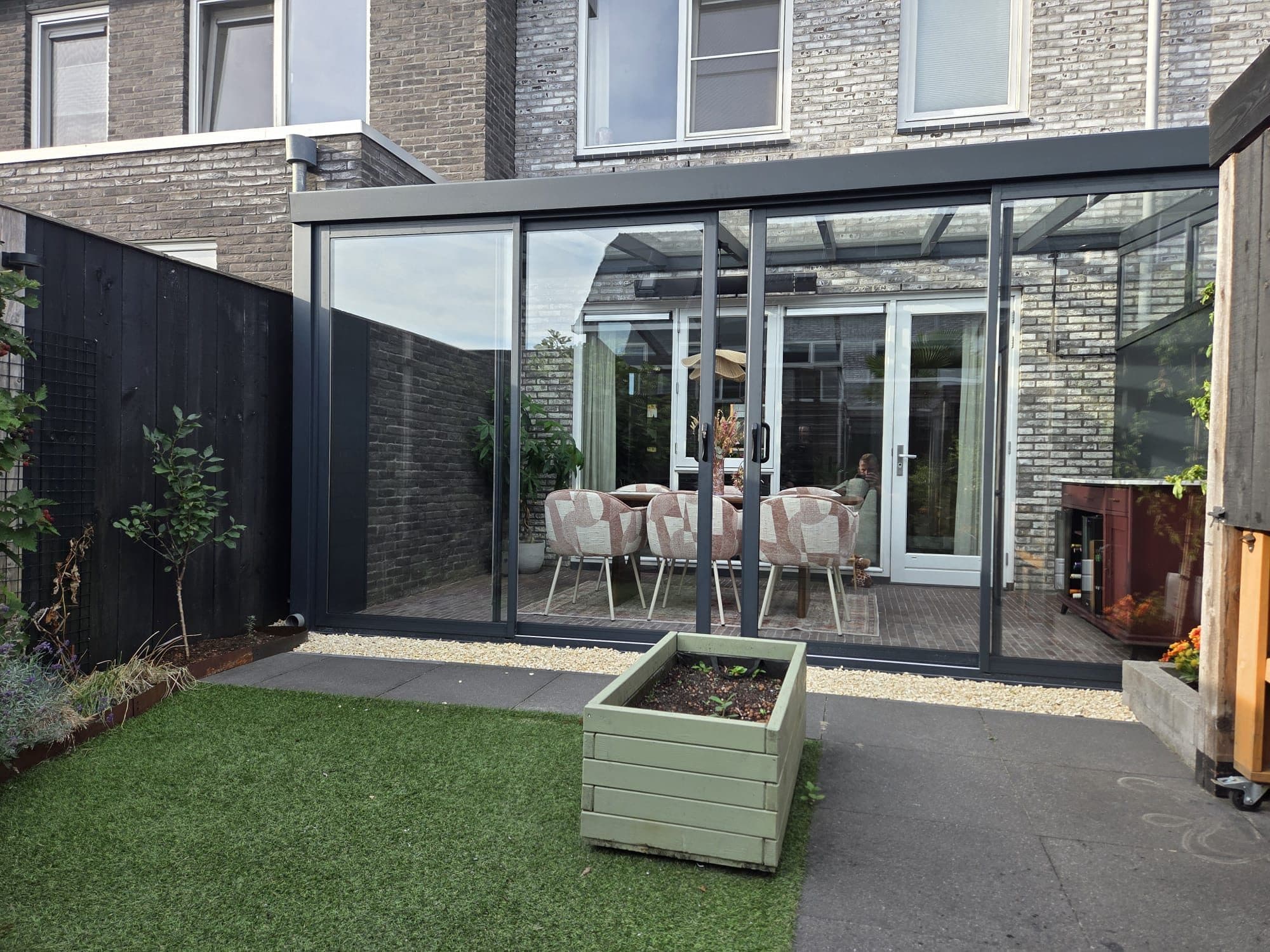 Antraciet veranda met glazen schuifwanden achter een rijwoning, gezien vanaf een tuin met kunstgras en een plantenbak