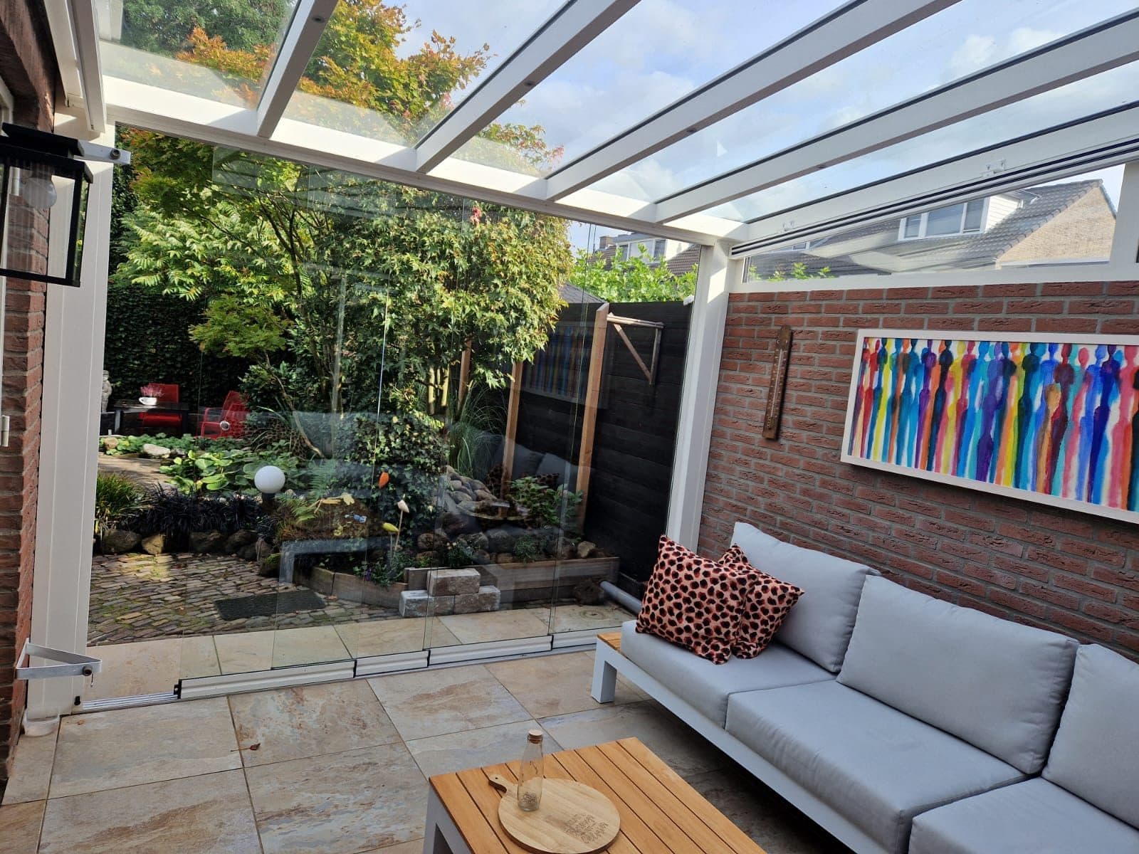 Witte veranda met glasdak en een grijze loungebank tegen een bakstenen muur, met zicht op een weelderige tuin