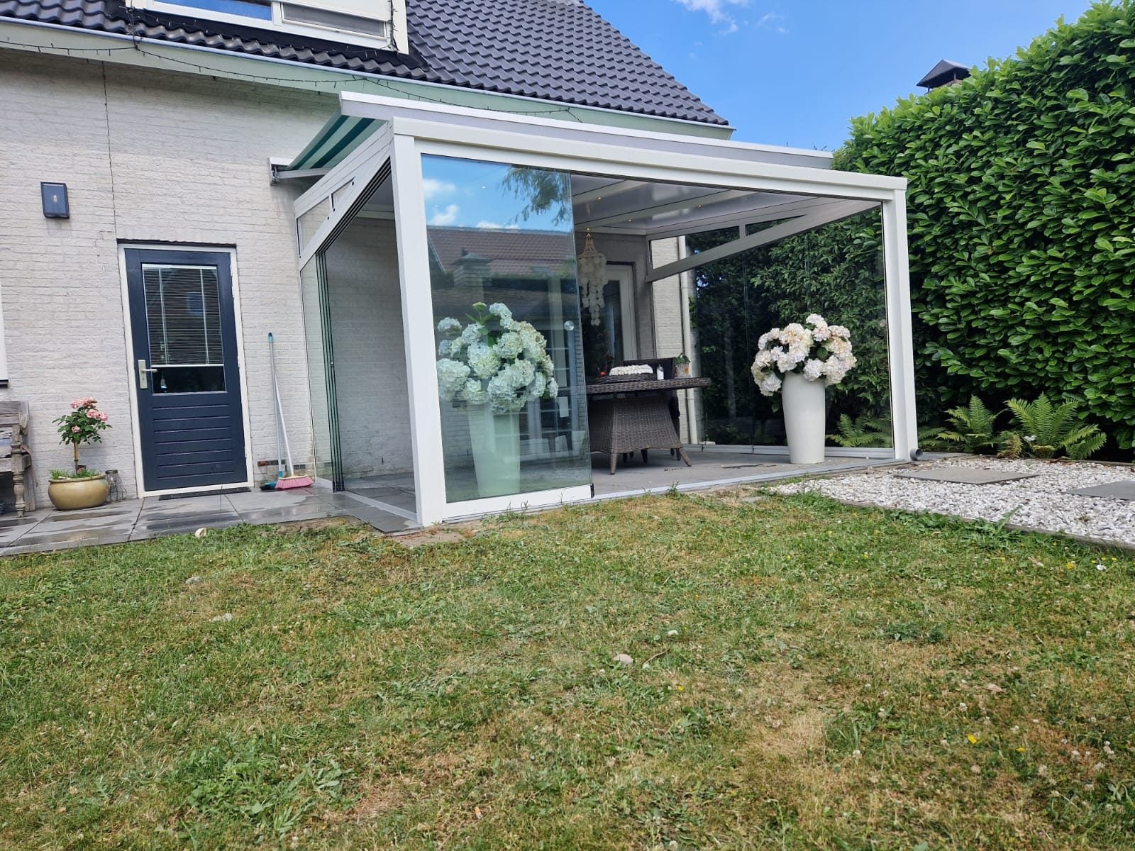 Witte veranda met glazen wanden en witte hortensia's in potten, aan een witte woning met een hoge haag ernaast