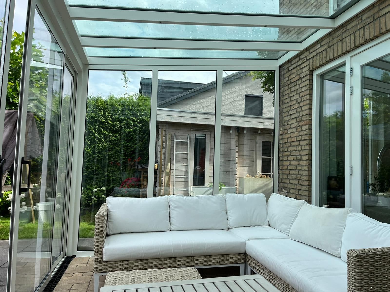 Witte loungebank onder een witte veranda met glasdak en glazen schuifwanden, met zicht op de tuin
