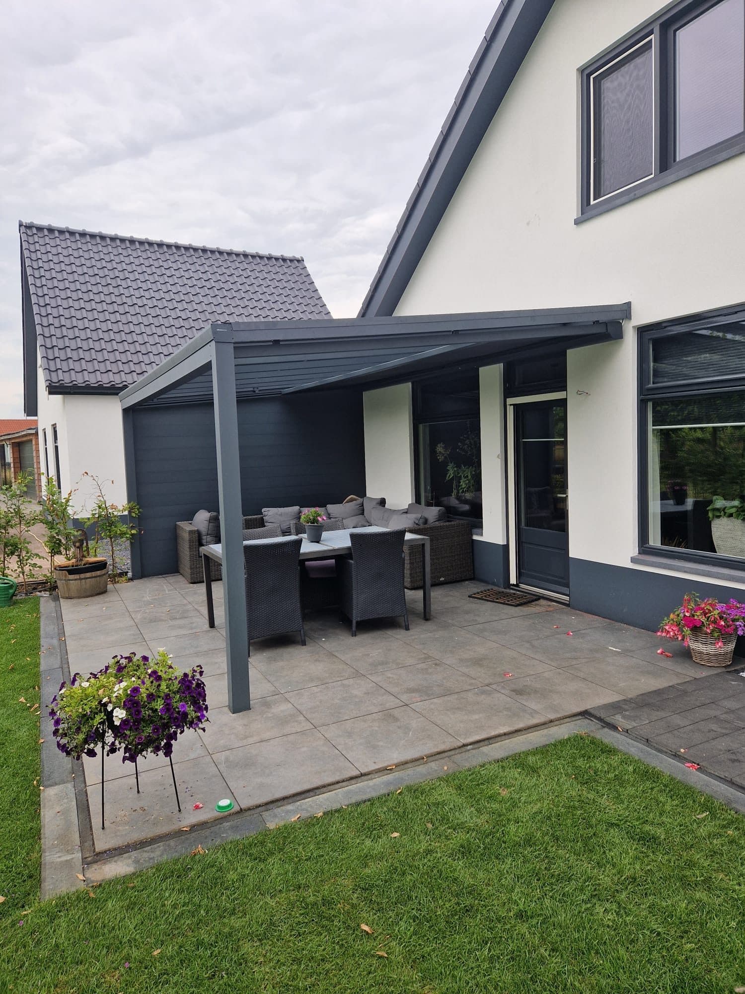Antraciet overkapping met een dichte zijwand aan een witte woning, boven een terras met loungeset en eettafel