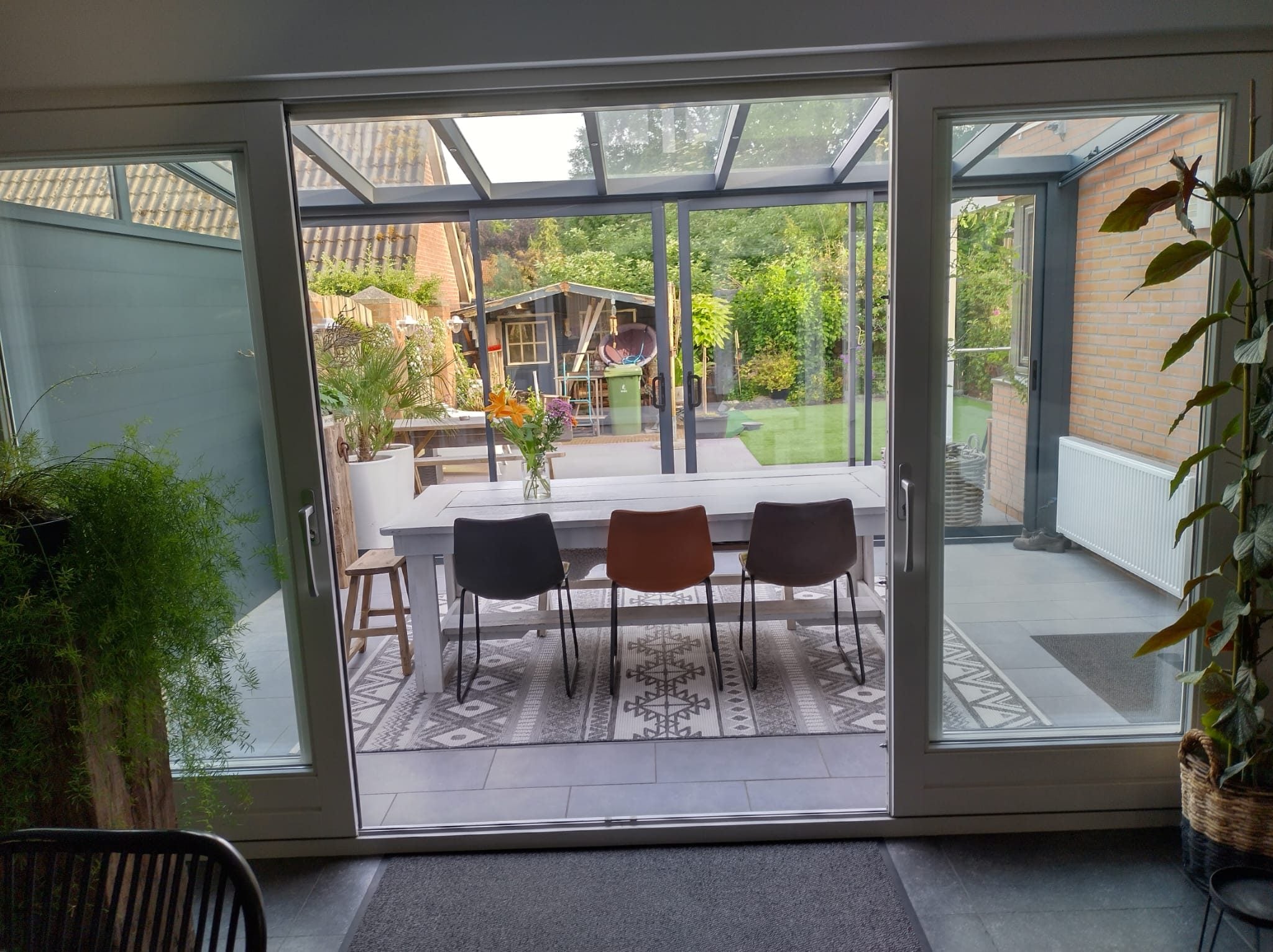 Zicht vanuit de woning door een open schuifpui naar een veranda met glasdak en eettafel, met de tuin erachter