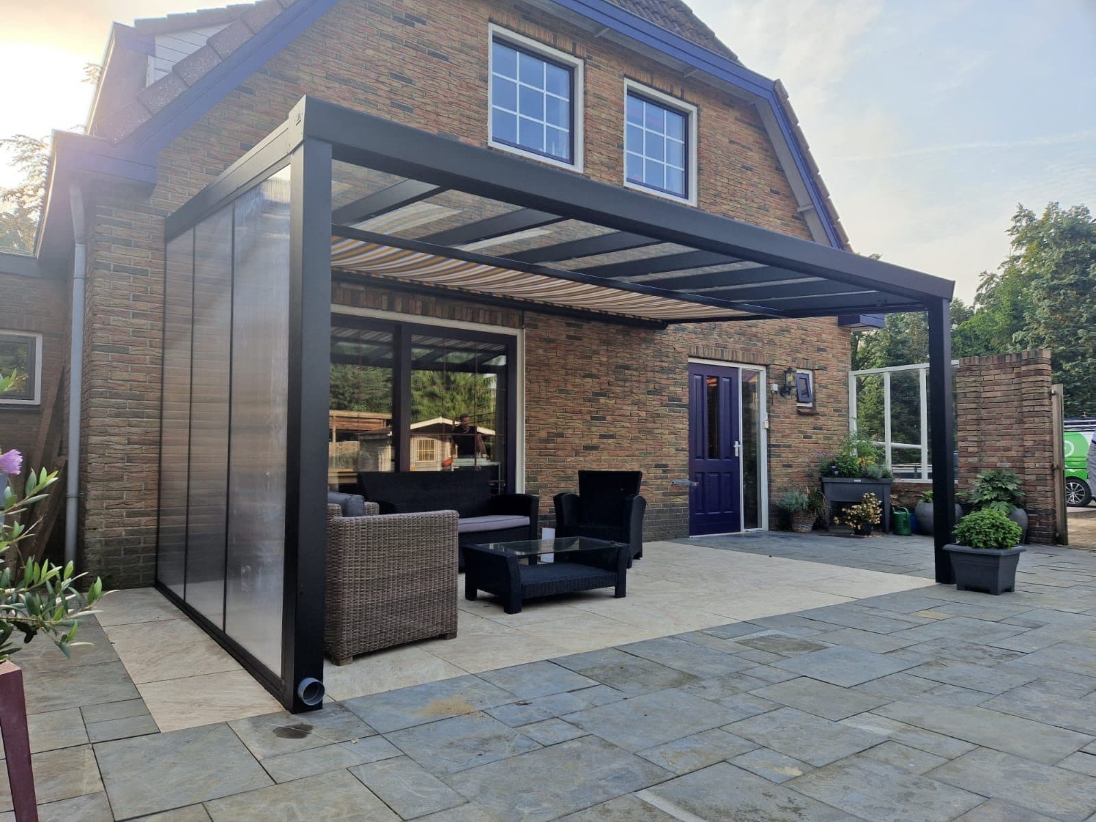 Antraciet veranda met een lichtdoorlatende zijwand en gestreepte zonwering onder het dak, boven een loungeset op een natuurstenen terras