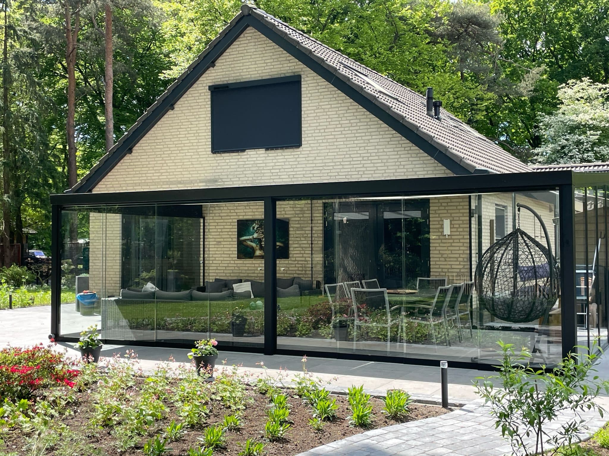 Zwarte veranda met rondom glazen schuifwanden en een hangstoel, aan een woning tussen hoge bomen
