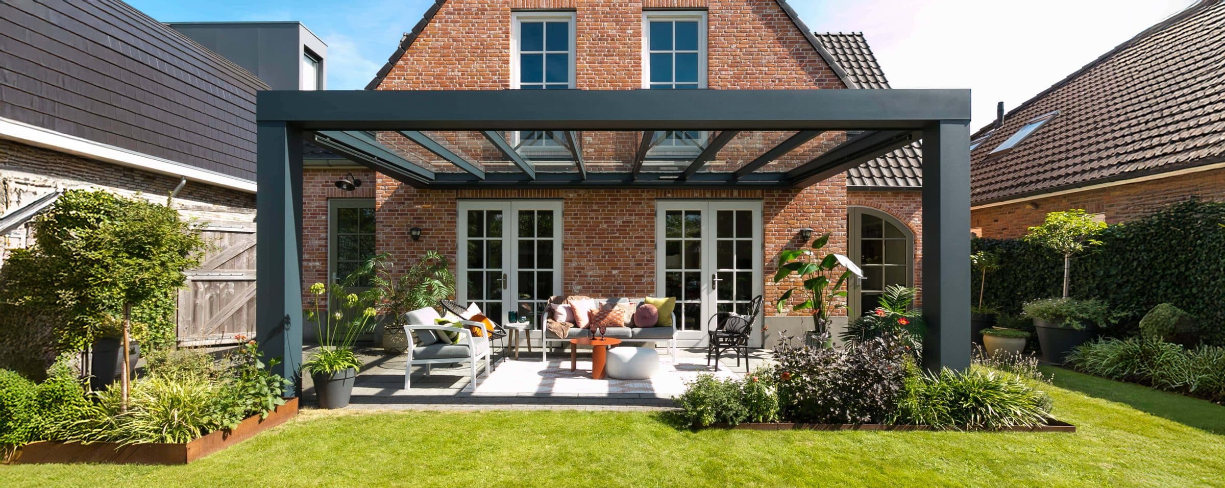 Antraciet overkapping in kubusvorm met dikke vlakke goot, twee staanders en glasdak voor een bakstenen woning, met loungeset in de tuin