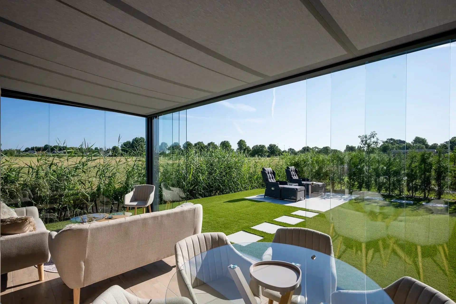 Loungebank en eettafel onder een vrijstaande overkapping met glazen wanden en uitzicht over weilanden