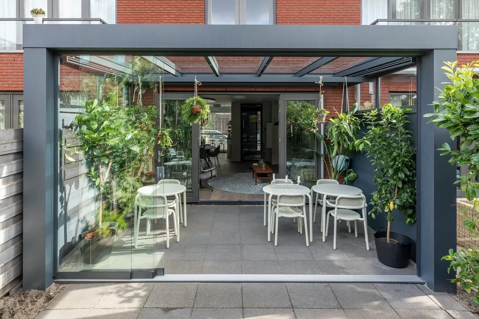 Antraciet Cube-overkapping met glazen wanden achter een rijwoning, vol hangplanten, met witte tafels en zicht door naar de woonkamer