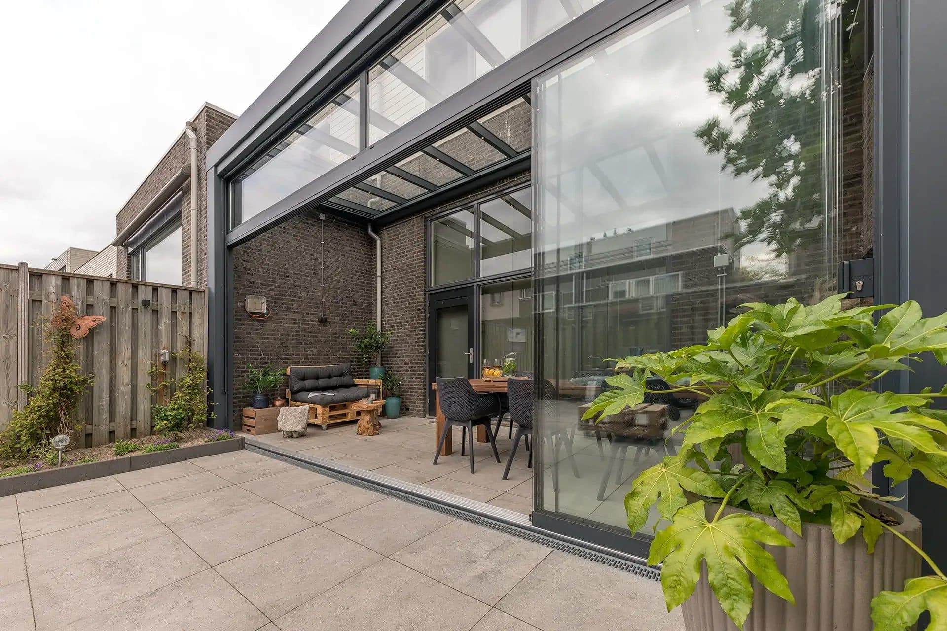Antraciet overkapping met glasdak en glazen wanden achter een rijwoning, gezien vanaf een licht tegelterras met een grote plant