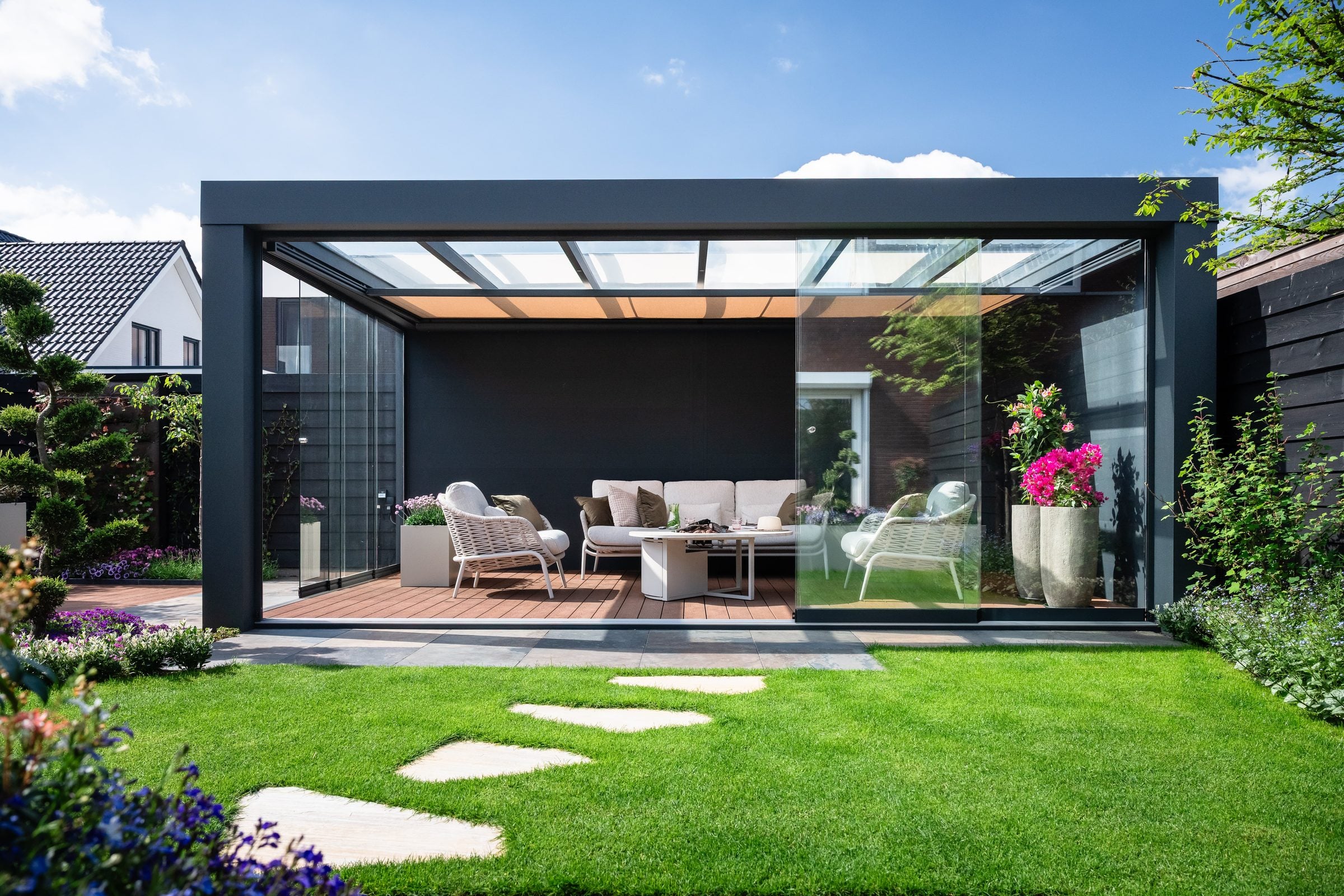 Vrijstaande antraciet Cube met glasdak en glazen schuifwanden achter in een tuin, met een loungeset op een houten vloer en stapstenen in het gazon