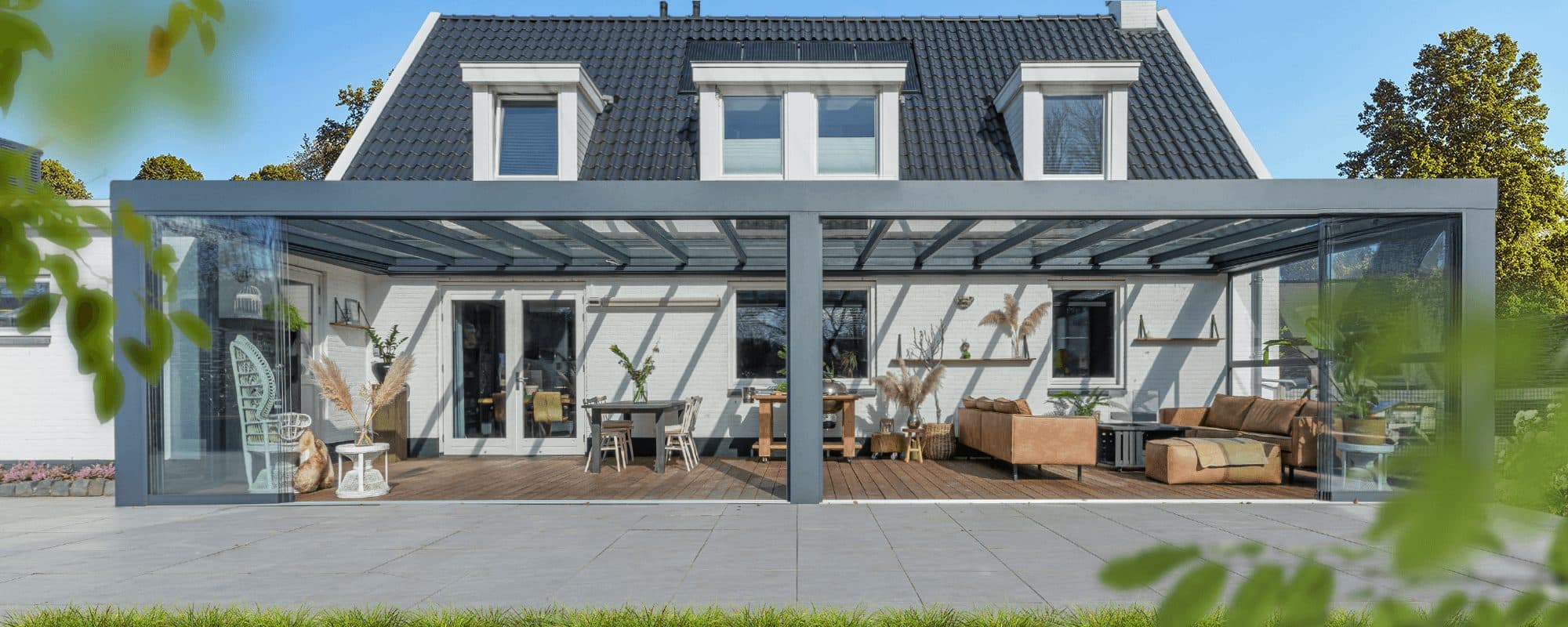 Brede antraciet overkapping met vlak dak en glazen wanden over de hele achtergevel van een witte woning met dakkapellen, met roze bloemen op de voorgrond