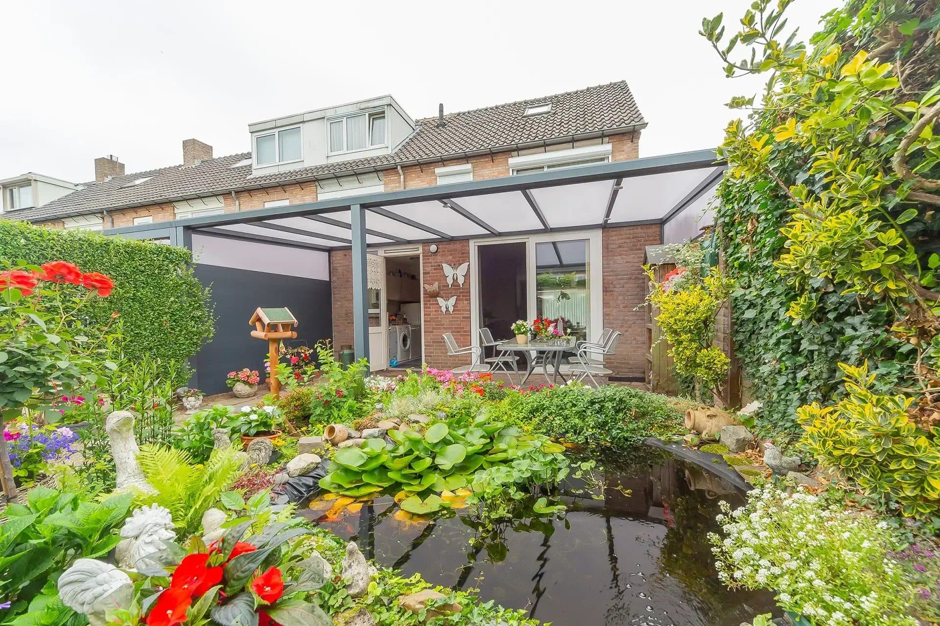 Antraciet veranda met polycarbonaat dak achter een rijwoning, gezien over een vijver en een bloeiende tuin vol kleur