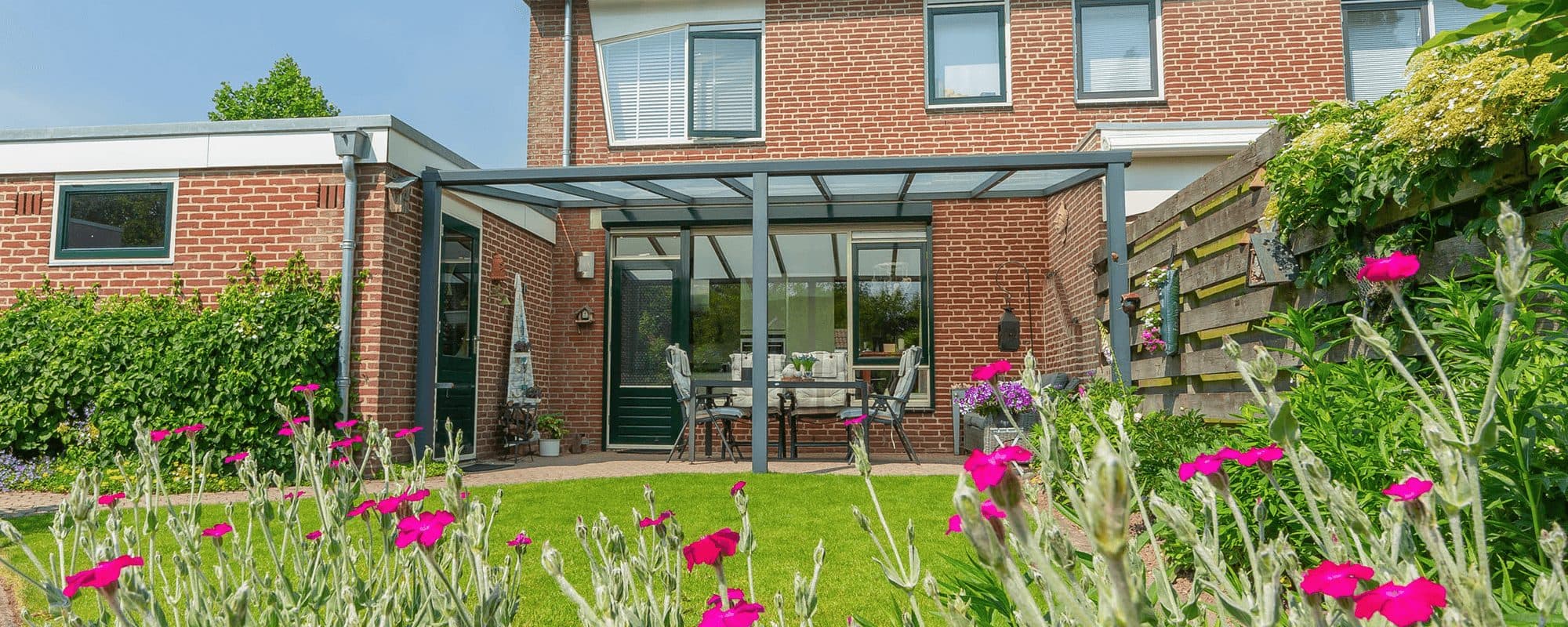 Antraciet veranda met glasdak achter een bakstenen woning, gezien over een gazon met bloeiende planten