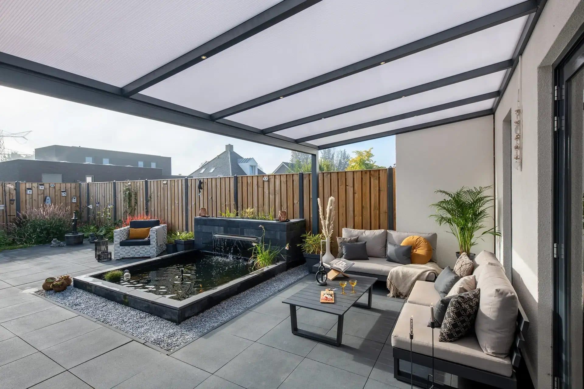 Loungebank en fauteuils onder een antraciet veranda met polycarbonaat dak, naast een vijver op een tegelterras