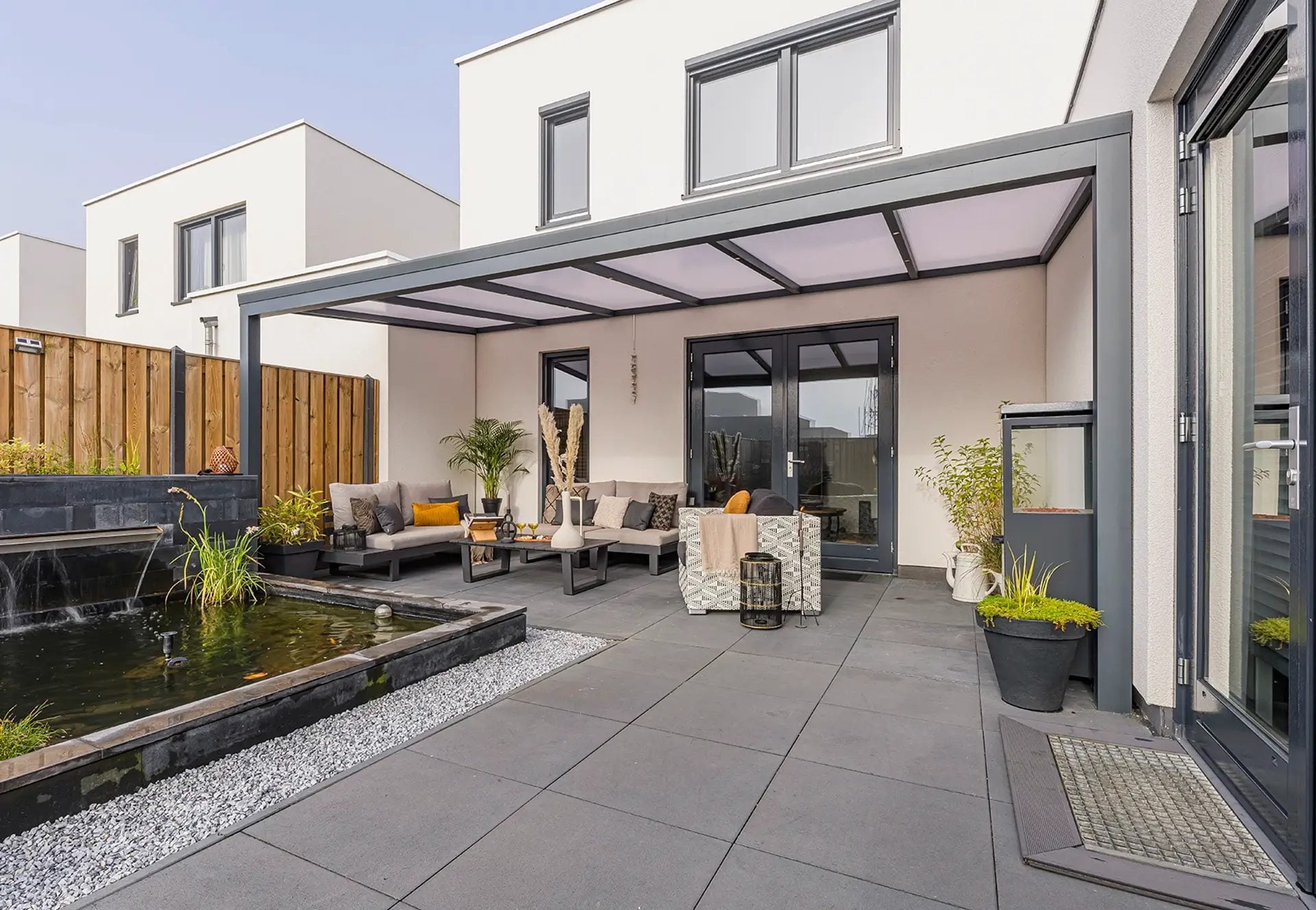 Antraciet veranda met opaal polycarbonaat dak boven een terras met loungeset en een vijver, tegen een witte woning