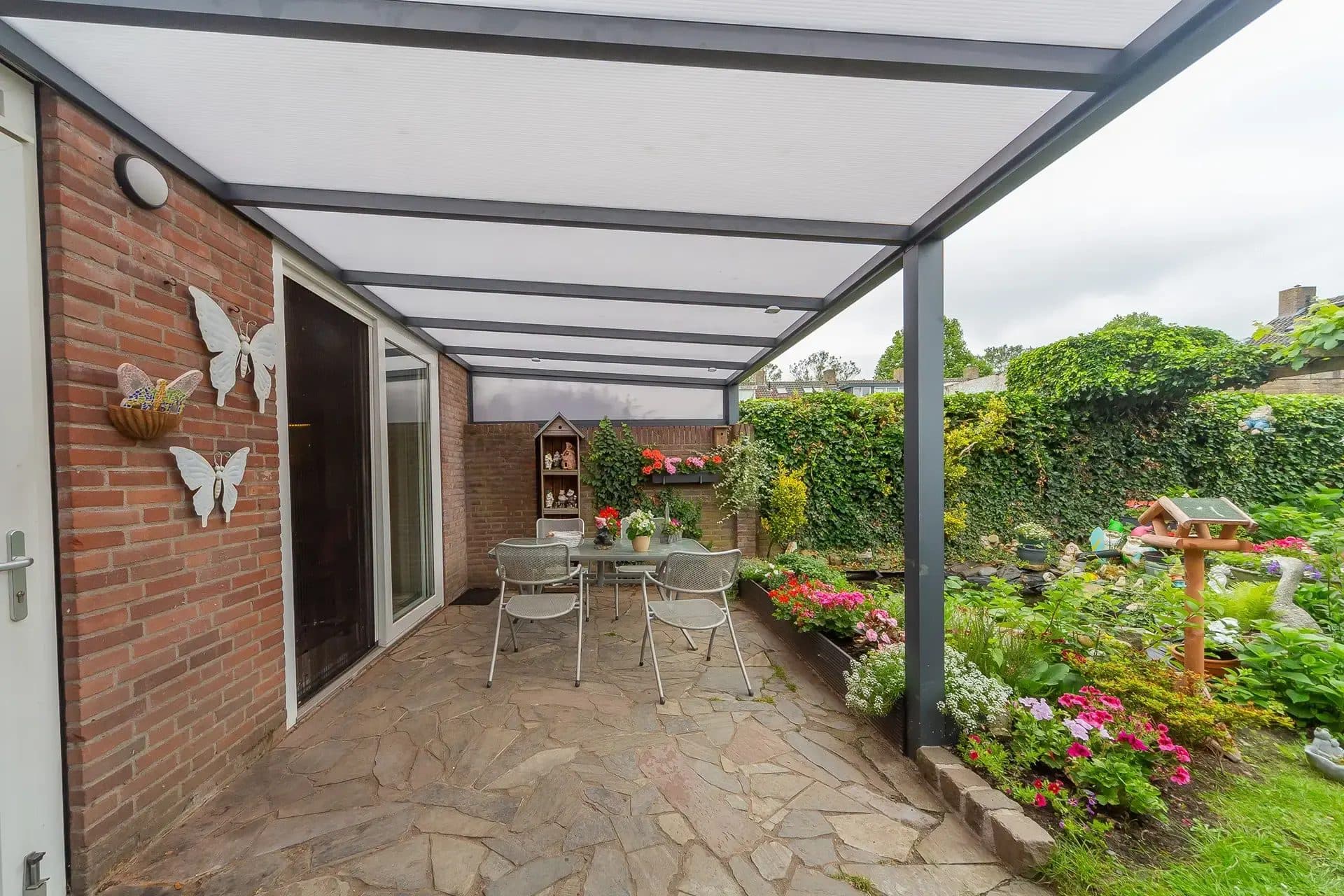Tuinset op een terras van natuursteen onder een antraciet veranda met polycarbonaat dak, met zicht op een bloeiende tuin