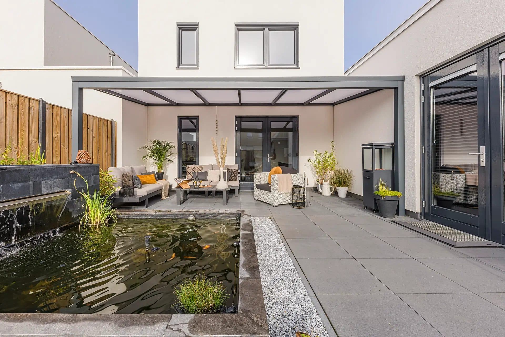 Antraciet veranda met opaal polycarbonaat dak tegen een witte woning, met loungeset op het terras en een vijver op de voorgrond
