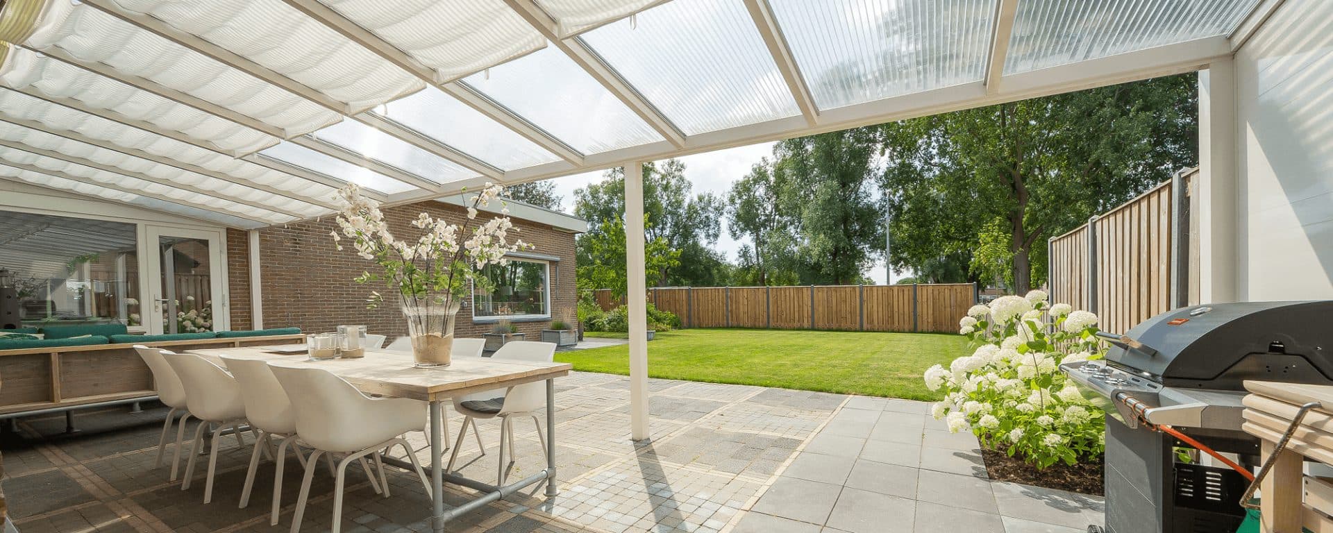 Witte veranda met lichtdoorlatend dak en doek eronder, boven een lange eettafel, met witte hortensia's en een barbecue en zicht op het gazon