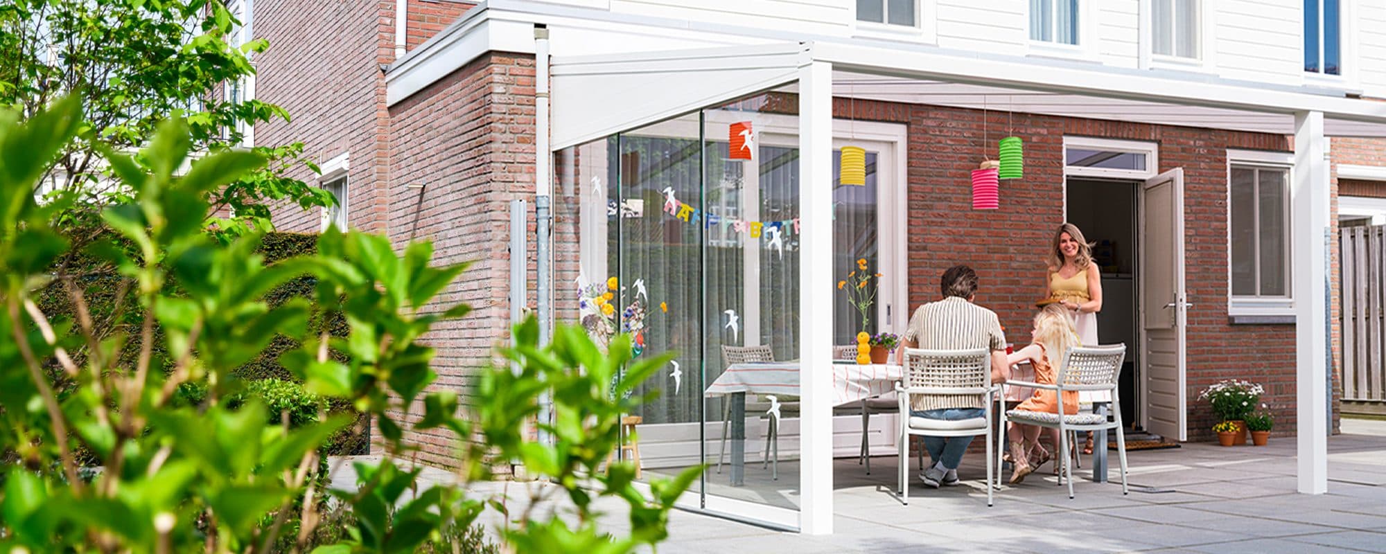 Witte veranda met glazen schuifwanden tegen een bakstenen woning, met een gezin aan tafel en feestversiering