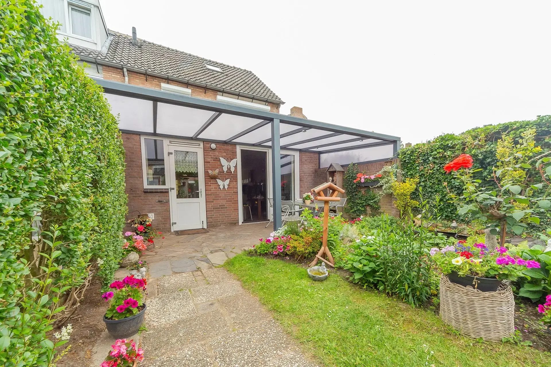 Antraciet veranda met polycarbonaat dak en zichtbare dakhelling tegen een bakstenen rijwoning, met een tuinpad en bloeiende planten
