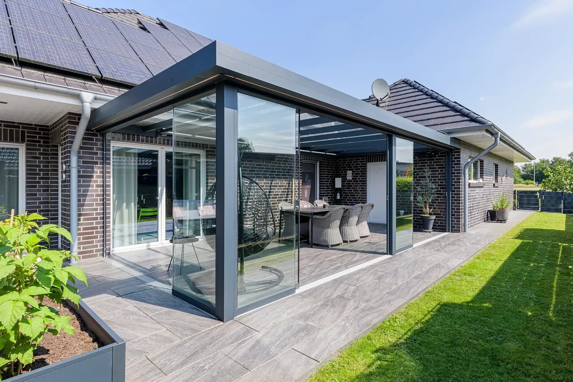 Antraciet veranda met vlak dak en glazen wanden in de hoek van een bungalow, met eethoek en een smal gazon langs het terras