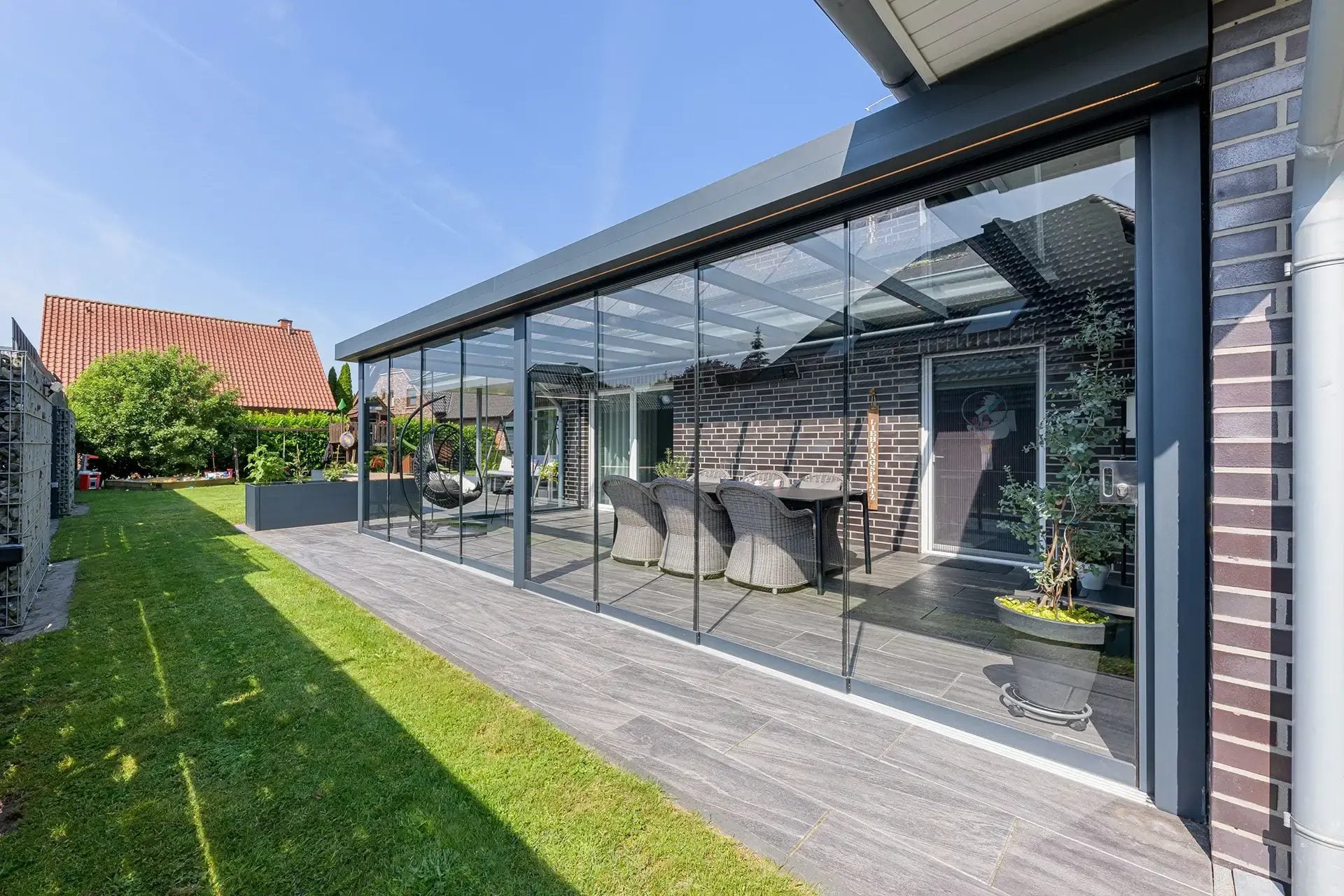 Lange antraciet veranda met vlak dak en glazen wanden langs een gazon, gezien vanaf de tuin