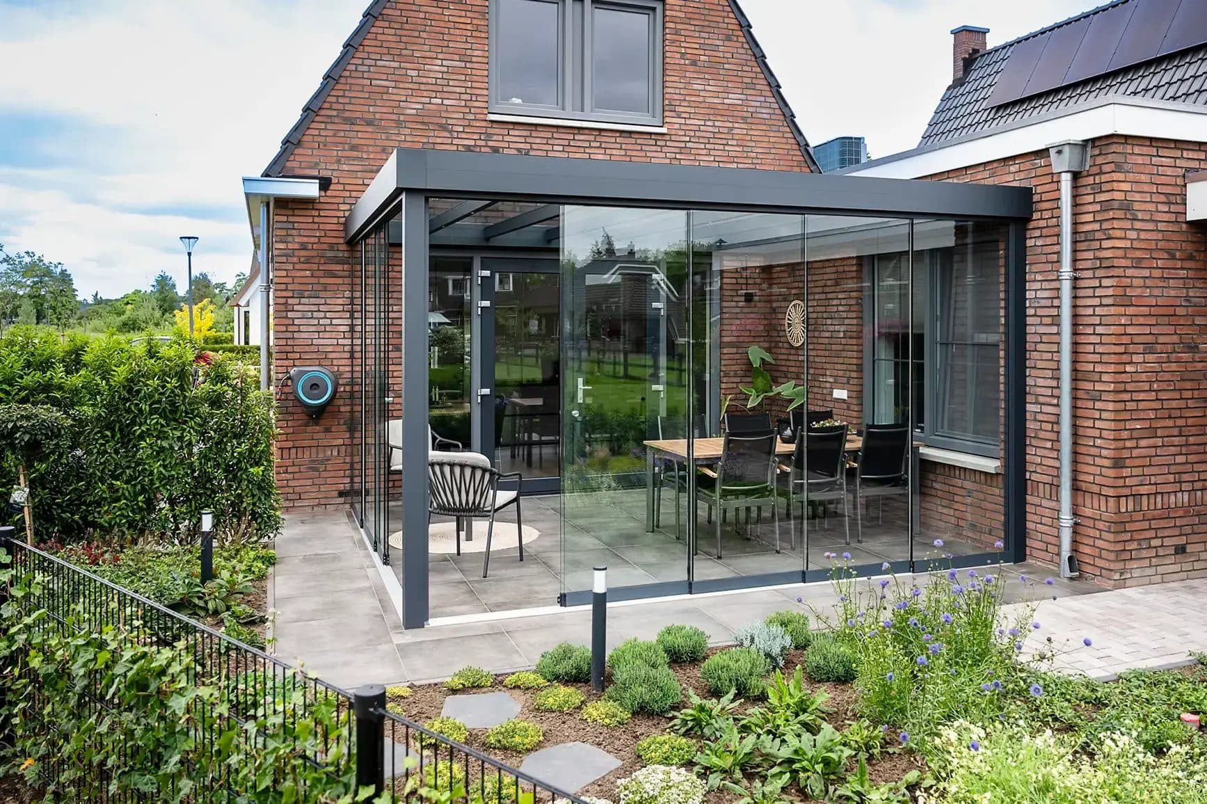 Antraciet veranda met vlak dak en rondom glazen wanden tegen een bakstenen woning met puntgevel, met eettafel binnen en een border ervoor