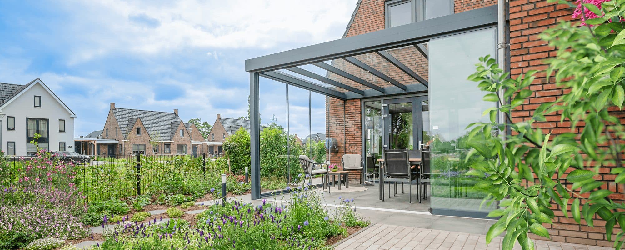 Antraciet veranda met vlak glasdak en glazen wanden aan een bakstenen woning, gezien vanuit een tuin met bloeiende borders