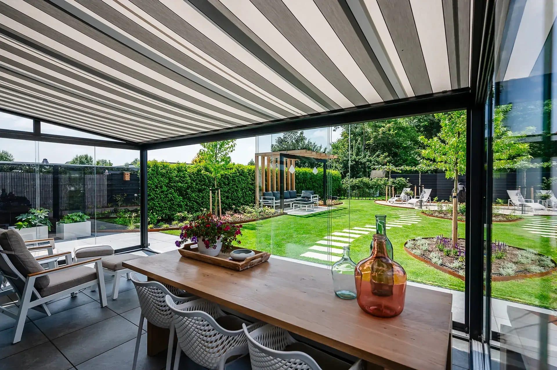 Eettafel onder een veranda met gestreept doek onder het glasdak en glazen wanden, met uitzicht over een grote tuin