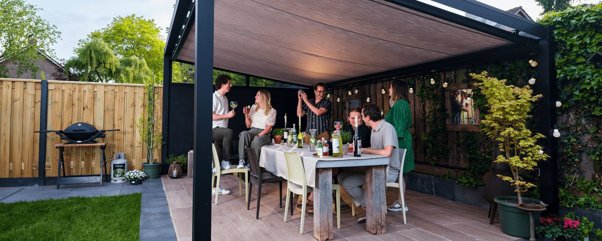 Gezelschap aan een gedekte tafel onder een antraciet overkapping met doek onder het dak en lichtslingers, met barbecue op het gazon