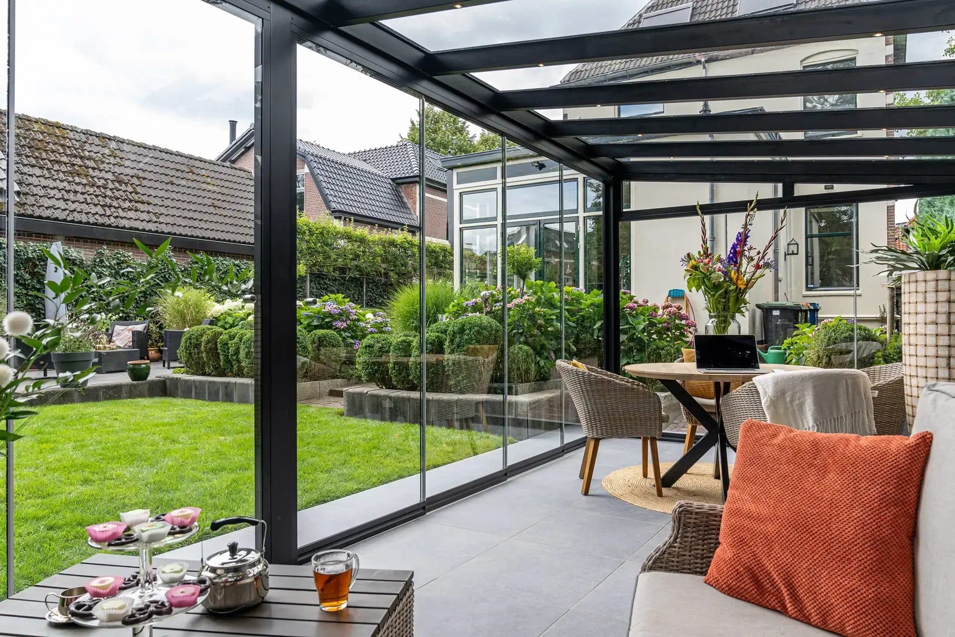 Thee en gebak op een tafeltje onder een zwarte overkapping met glazen wanden, met zicht op een eethoek en een groene tuin