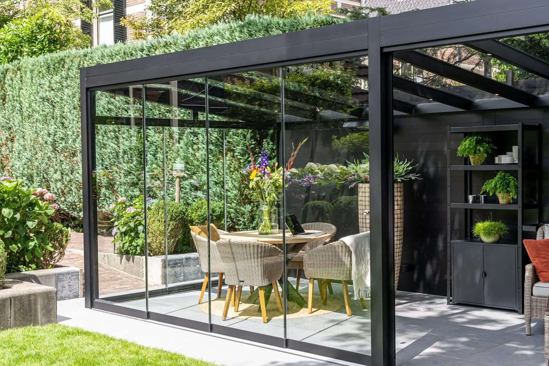 Zwarte vrijstaande overkapping met glasdak en glazen schuifwanden tegen een hoge haag, met eettafel, bloemen en een open kast