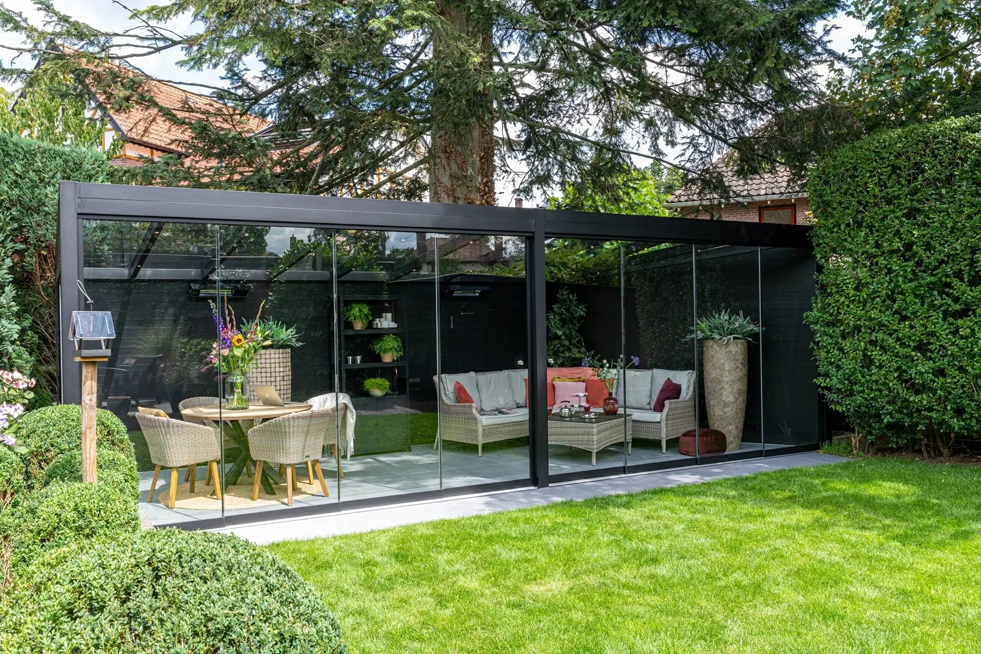 Zwarte vrijstaande overkapping met glasdak en glazen schuifwanden in een tuin met hagen, met eethoek en loungeset