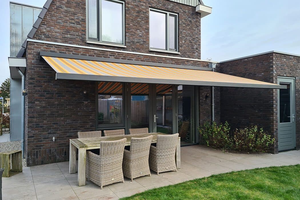 Uitgerold knikarmscherm met geel-grijs gestreept doek boven een terras met tuinset