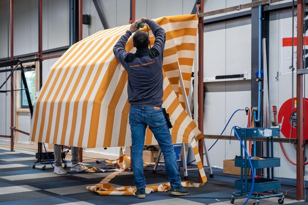 Medewerker spant een oranje-wit gestreept doek over het frame van een markies in de werkplaats