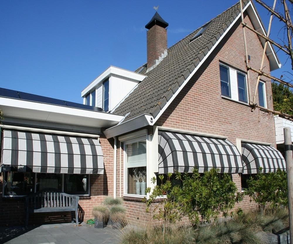 Bakstenen woning met zwart-wit gestreepte markiezen boven de ramen, met een tuin met siergrassen ervoor