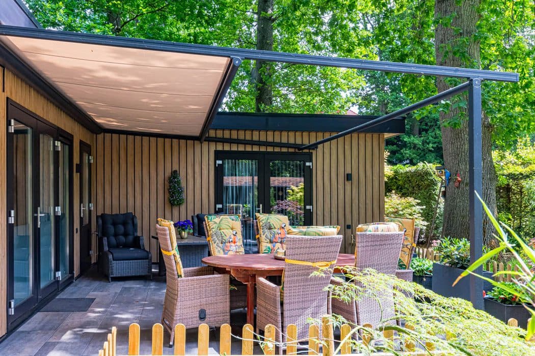 Pergolazonwering met zandkleurig doek tussen een woning met houten gevel en een losse staander, boven een tuinset