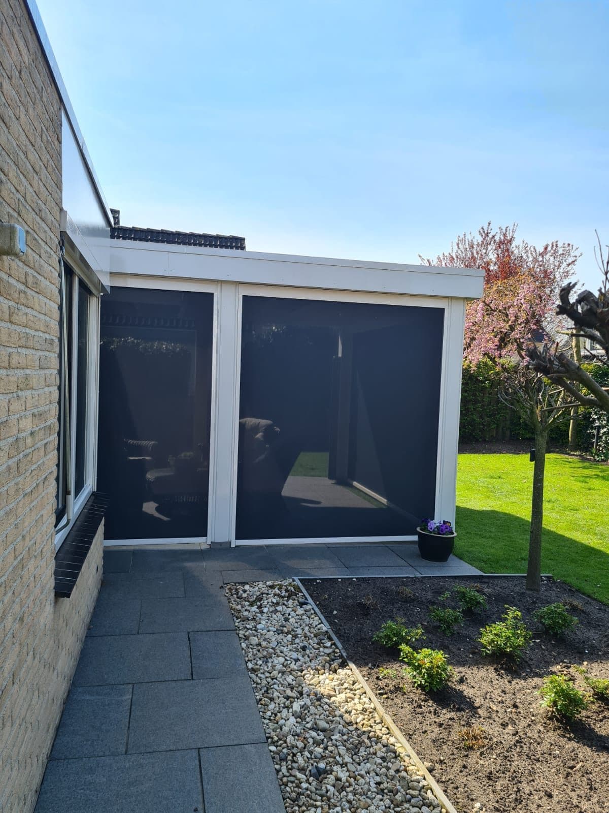 Twee neergelaten screens in de open zijkant van een witte aanbouw, met een nieuwe border ervoor