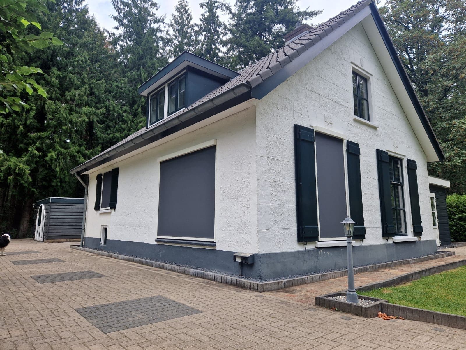 Antraciet screens en groene luiken aan een witte woning tussen hoge naaldbomen