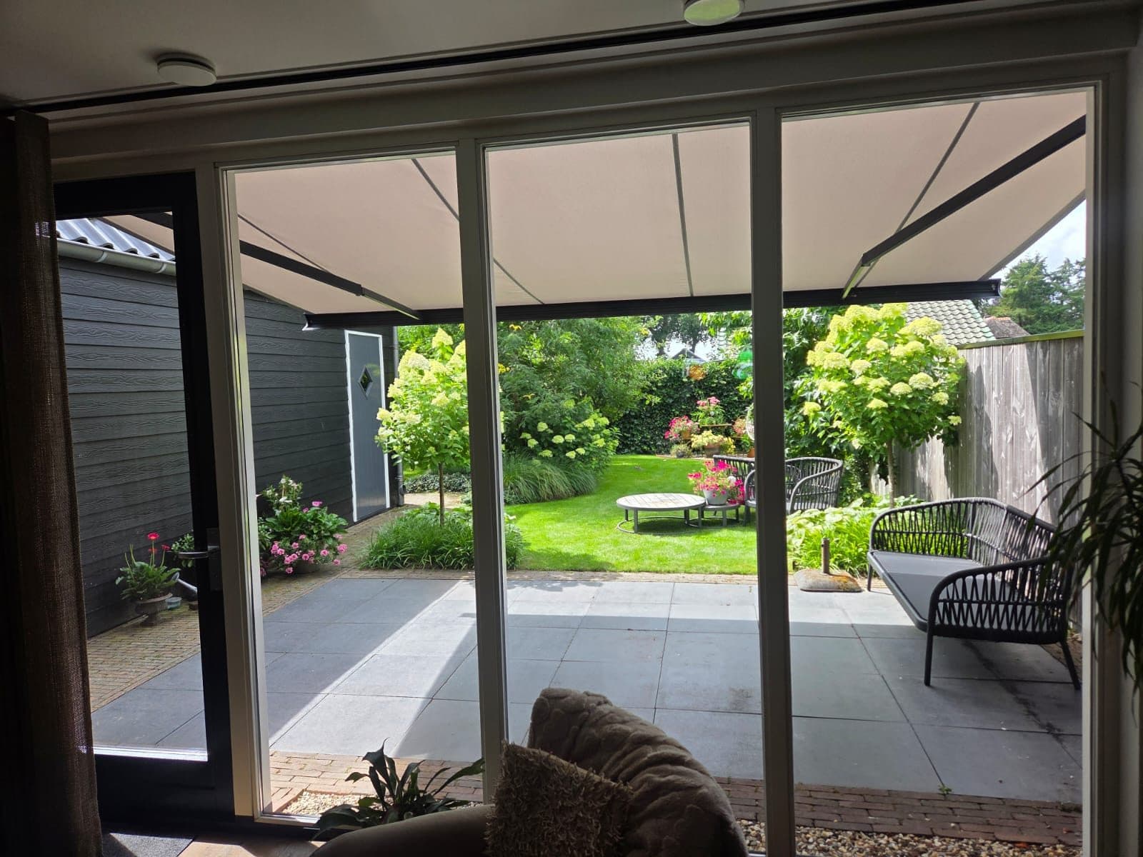 Zicht vanuit de woning onder een uitgerold terrasscherm door naar een bloeiende tuin met hortensia's