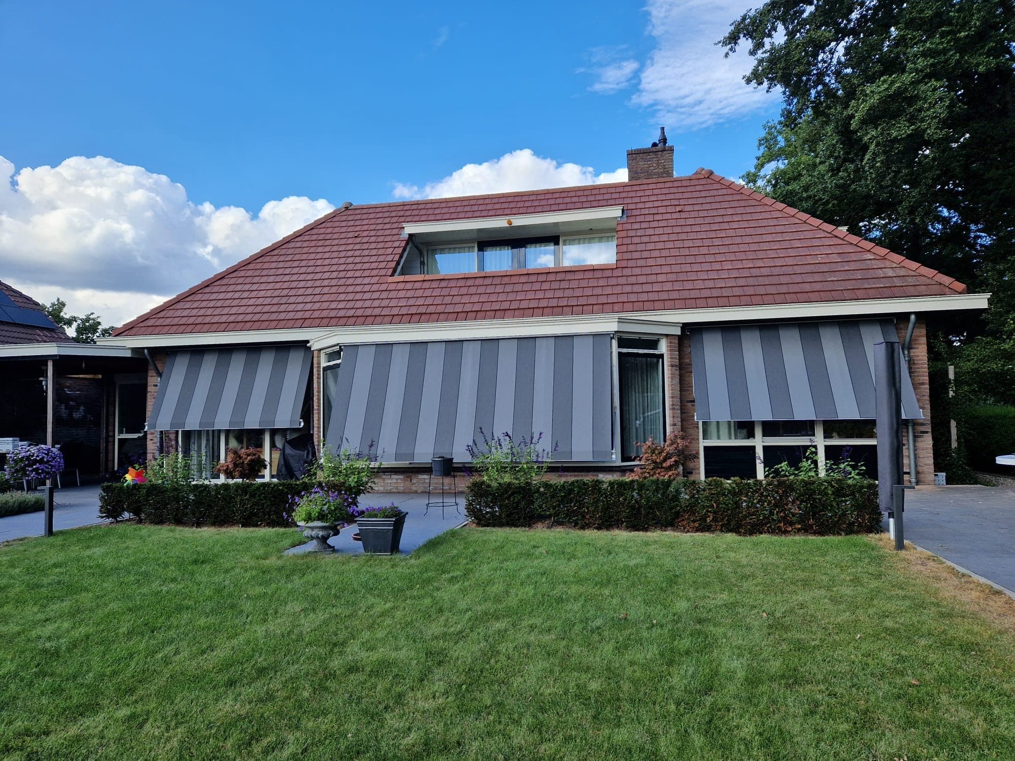 Grijs gestreepte zonneschermen boven de ramen van een bungalow met rood pannendak, gezien over een gazon