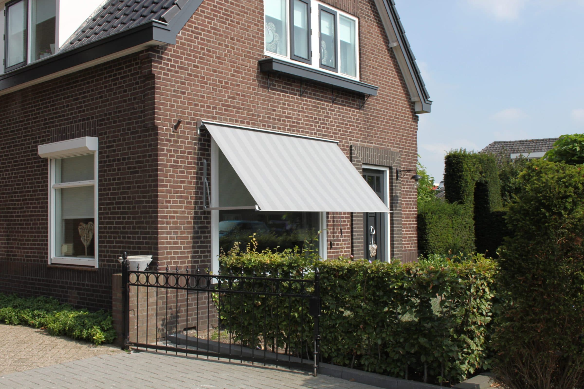 Bakstenen hoekwoning met een uitgeklapt uitvalscherm boven het raam naast de voordeur, achter een haag en een smeedijzeren hek