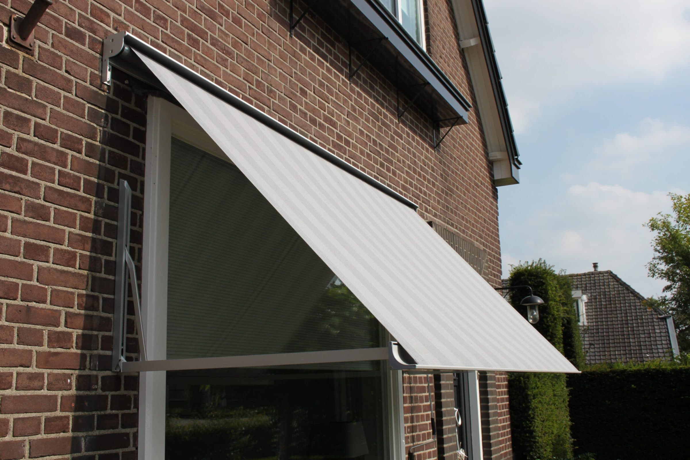 Uitvalscherm met lichtgrijs gestreept doek, schuin naar buiten voor een groot raam in een bakstenen gevel