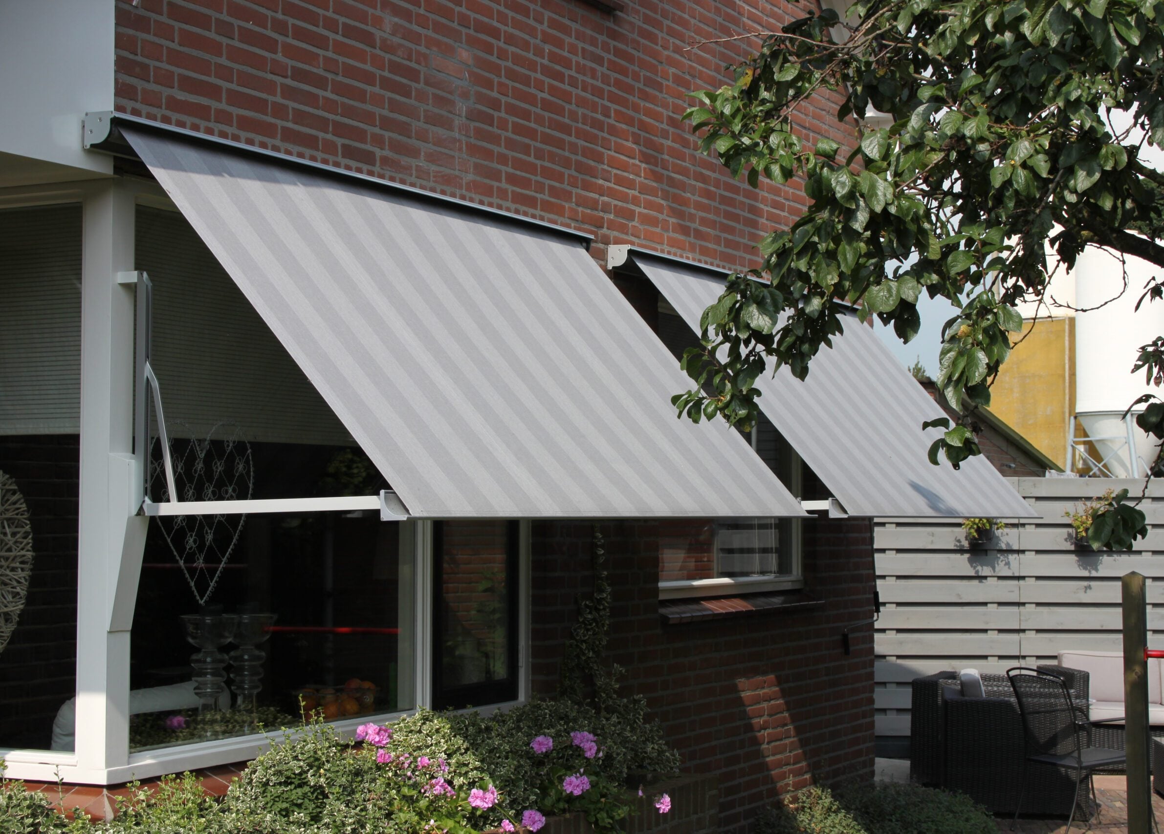 Twee uitvalschermen met lichtgrijs gestreept doek voor de ramen van een bakstenen woning, met een tuin met loungeset en bloemen ervoor