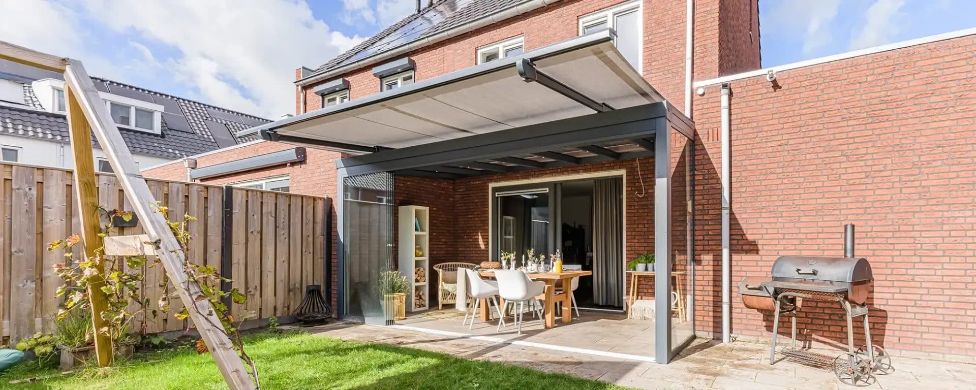 Antraciet veranda met bovendakzonwering achter een bakstenen rijwoning, met eettafel, barbecue en gazon