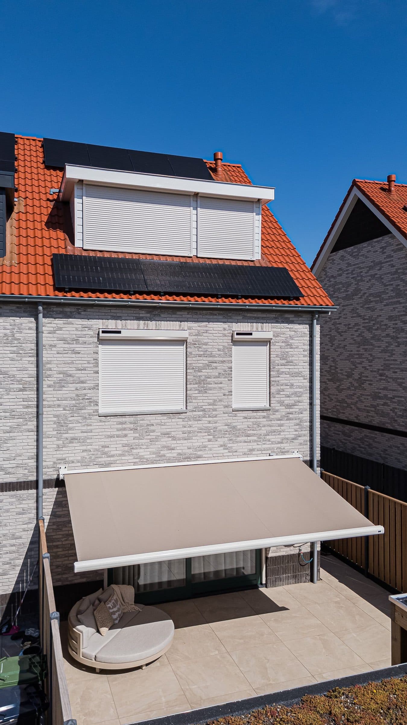 Achtergevel met een uitgerold knikarmscherm boven het terras en witte rolluiken op de verdieping