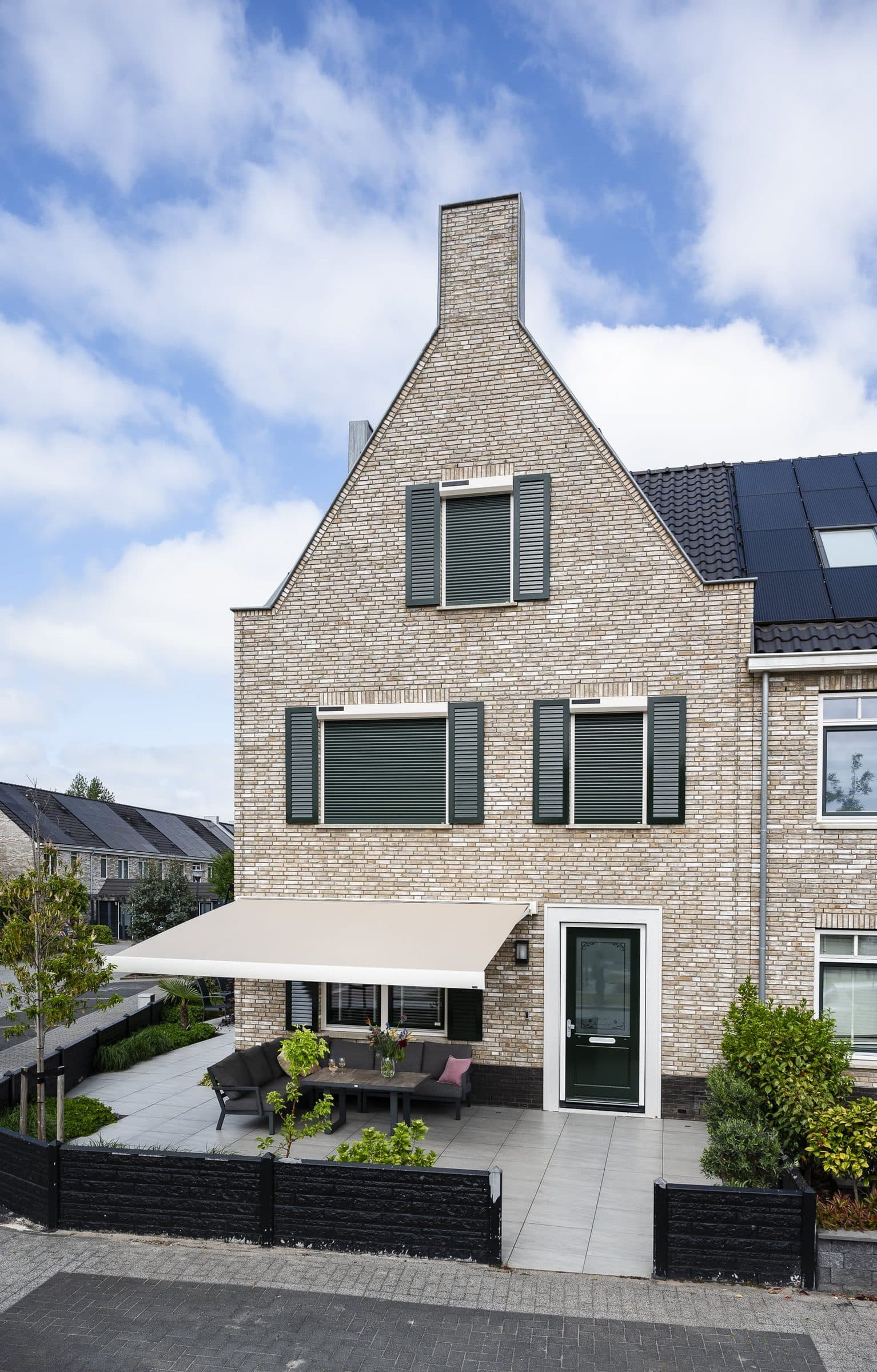 Uitgerold knikarmscherm boven het terras van een bakstenen hoekwoning met luiken en een puntgevel