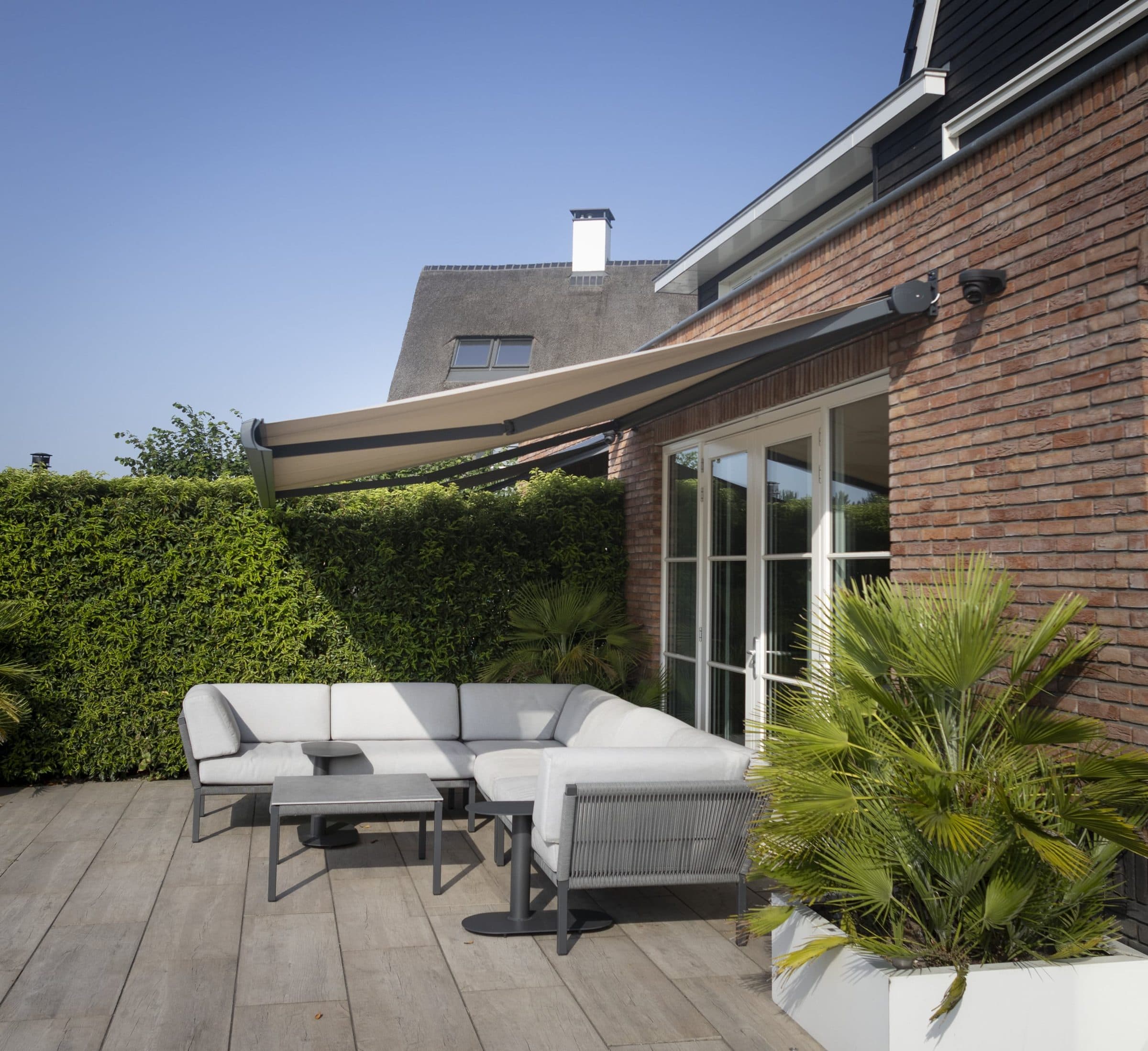 Uitgerold beige knikarmscherm boven een witte loungeset op het terras van een bakstenen woning