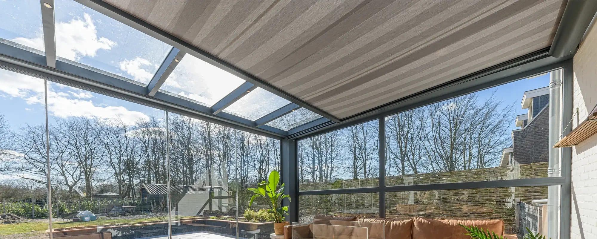 Grijze onderdakzonwering onder het glasdak van een veranda met glazen wanden en een loungebank