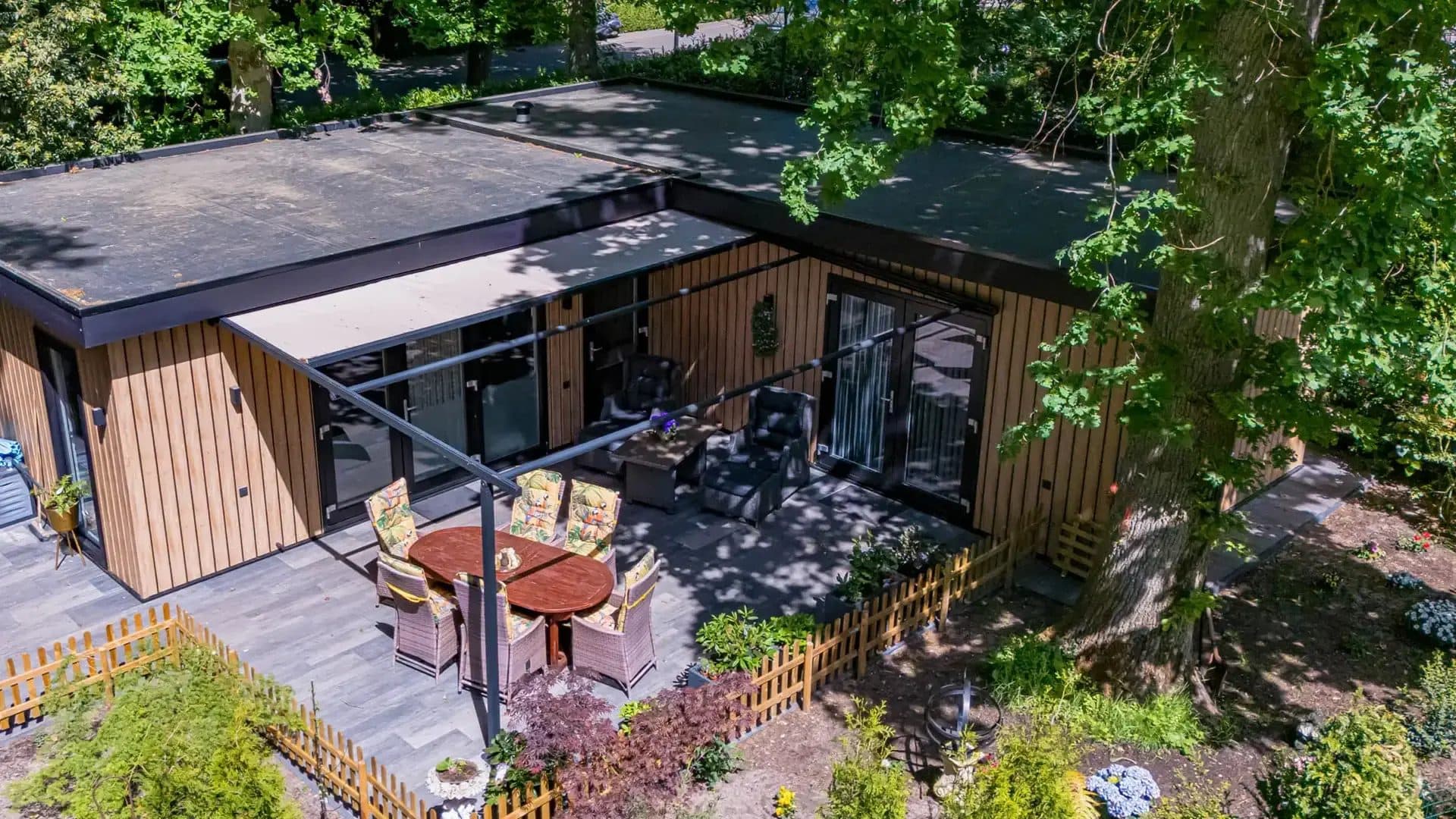 Pergola met doek naast een houten tuinhuis met eettafel en rieten stoelen, van bovenaf gezien tussen de bomen